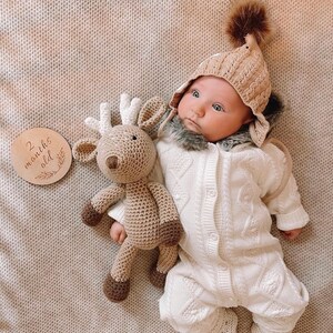 May include: A baby in a beige knitted hat with a brown pom-pom and a white knitted jumpsuit. A crocheted reindeer toy and a wooden disc that says "2 months old" are also in the image.