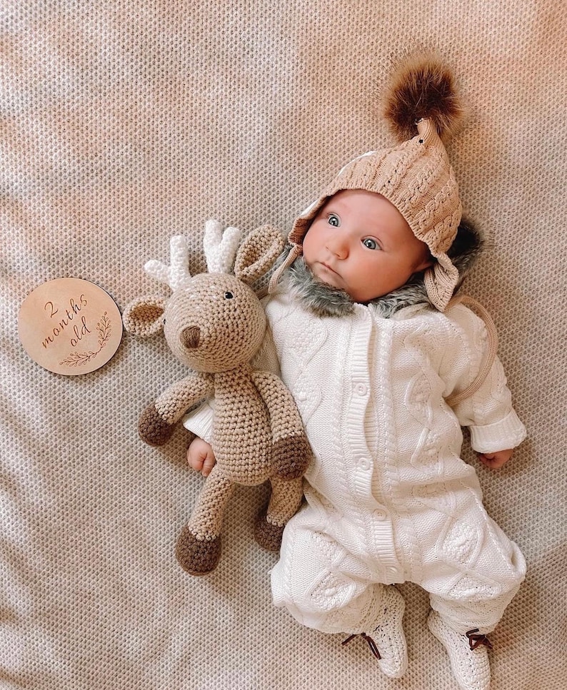 May include: A baby in a beige knitted hat with a brown pom-pom and a white knitted jumpsuit. A crocheted reindeer toy and a wooden disc that says "2 months old" are also in the image.