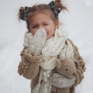 May include: A child wearing a beige knitted cardigan, mittens, and a white scarf, with a crocheted deer toy. The child has hair bows. The background is a snowy scene, typical of winter.