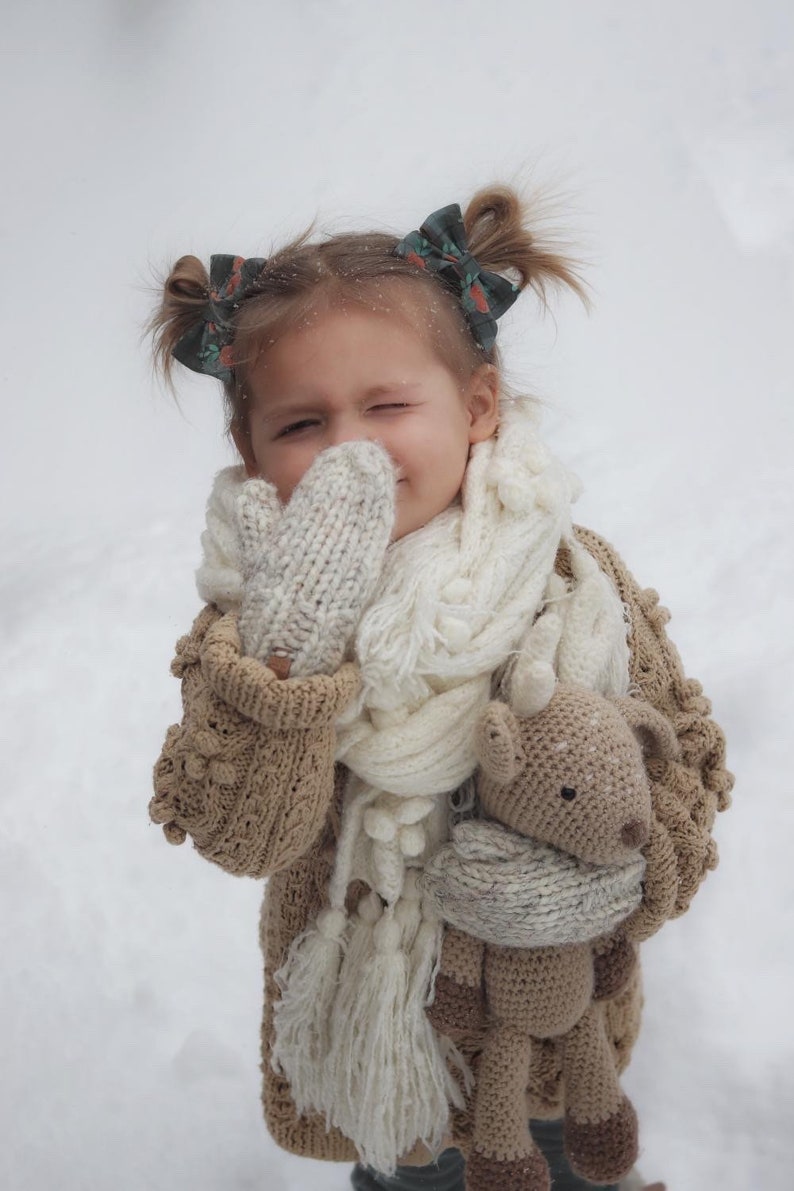 May include: A child wearing a beige knitted cardigan, mittens, and a white scarf, with a crocheted deer toy. The child has hair bows. The background is a snowy scene, typical of winter.