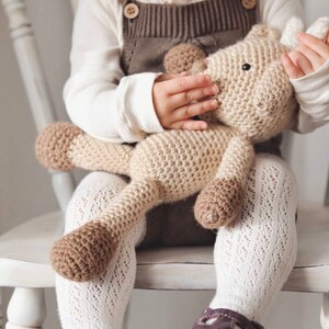 May include: A beige and brown crocheted giraffe toy. The giraffe has a long neck and four legs. A child is holding the toy, wearing a white long-sleeved top, brown dungarees, white patterned tights, and purple shoes.