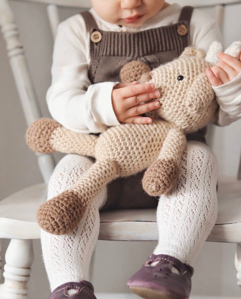 May include: A beige and brown crocheted giraffe toy. The giraffe has a long neck and four legs. A child is holding the toy, wearing a white long-sleeved top, brown dungarees, white patterned tights, and purple shoes.