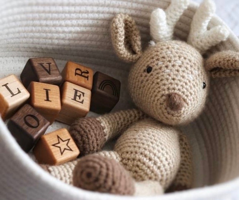 May include: A crocheted deer toy with a light brown body, white antlers, and dark brown accents sits in a woven basket. Wooden blocks spelling "OLIVER" and featuring a star and rainbow are scattered around the deer.