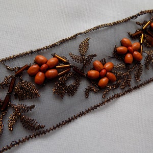 Pode incluir: Fita decorativa com um design floral. A fita tem uma base cinza transparente com detalhes de contas marrons. As flores são feitas de contas de madeira laranja e as folhas são de arame marrom.