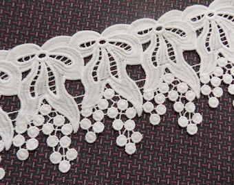 May include: White lace trim with a delicate floral design and small round beads. The trim features a repeating pattern of bows and clusters of beads.