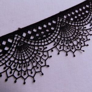 May include: Close-up of a black lace trim with a scalloped edge. The trim features a repeating pattern of openwork designs, including diamond shapes and fan-like motifs. The lace is set against a white background.