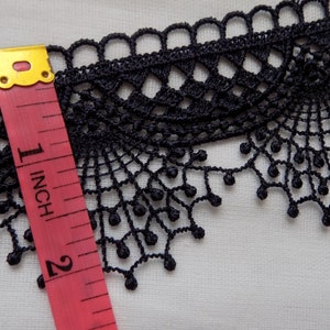 May include: Black lace trim with a scalloped edge and a delicate, openwork design. A pink and yellow measuring tape shows the trim is approximately 1 inch wide. The trim is on a white background.