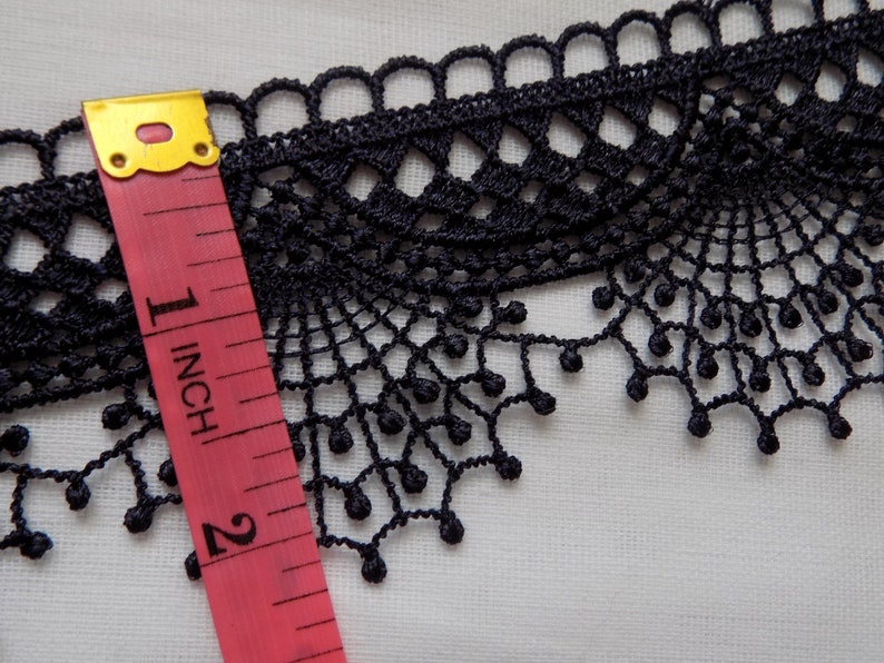 May include: Black lace trim with a scalloped edge and a delicate, openwork design. A pink and yellow measuring tape shows the trim is approximately 1 inch wide. The trim is on a white background.