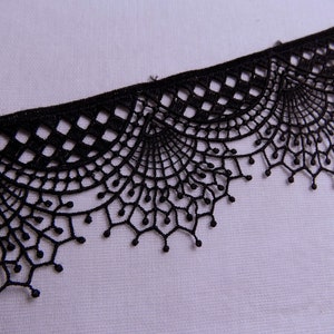 May include: Close-up of a black lace trim with a scalloped edge. The lace features a delicate, openwork design with geometric and floral patterns. The trim is likely used for sewing and crafting projects.