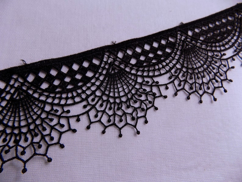 May include: Close-up of a black lace trim with a scalloped edge. The lace features a delicate, openwork design with geometric and floral patterns. The trim is likely used for sewing and crafting projects.