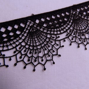 May include: Close-up of black lace trim with a scalloped edge. The lace features a geometric pattern at the top and a delicate, web-like design with small beaded details along the bottom edge. The trim is against a white background.