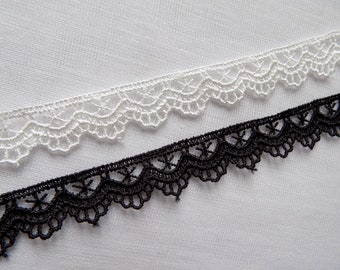 May include: Close-up of two decorative lace trims. One is white, the other is black. Both trims have a scalloped edge design. The lace is set against a white fabric background.