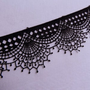 May include: Black lace trim with a delicate, scalloped edge. The trim features a repeating pattern of geometric shapes and fan-like designs. This decorative trim is suitable for various craft and sewing projects.