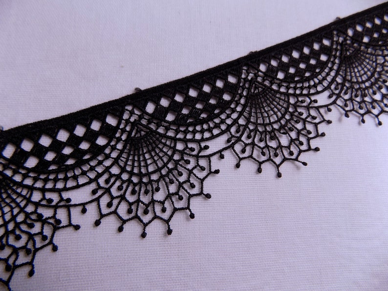 May include: Black lace trim with a delicate, scalloped edge. The trim features a repeating pattern of geometric shapes and fan-like designs. This decorative trim is suitable for various craft and sewing projects.