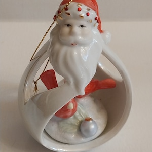 Op de afbeelding: Een keramische kerstversiering met de kerstman met een rode muts en baard, die een rode vogel vasthoudt. De versiering is wit met gouden accenten en een zilveren bal. Een gouden touwtje is bevestigd om op te hangen.