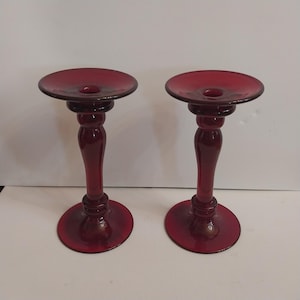 Może przedstawiać: Dwa rubinowoczerwone świeczniki szklane. Każdy ma okrągłą podstawę, smukłą, zakrzywioną nóżkę i płytką, okrągłą górę na świecę. Świeczniki tworzą pasujący zestaw.