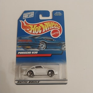 May include: A white Hot Wheels Porsche 930 die-cast toy car in its original blue blister packaging. The card features the Hot Wheels logo, the text Porsche 930, and the Mattel logo. The packaging includes the text Guaranteed For Life.