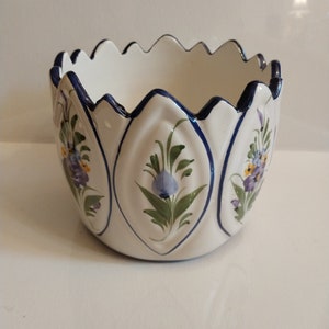 Pode incluir: Vaso de cerâmica branco com borda ondulada e acabamento azul. O vaso é decorado com desenhos florais pintados à mão em tons de azul, amarelo e roxo.