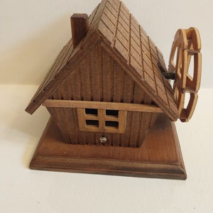 May include: Wooden music box shaped like a house with a small chimney and a spinning wheel on the side. The house has a small window with four panes.