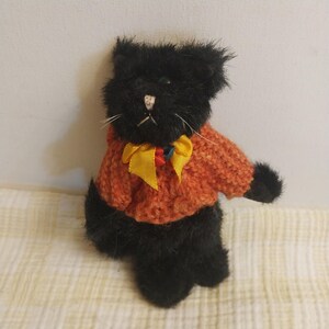May include: A black plush cat wearing an orange sweater with a yellow bow. The cat has white whiskers and a small pink nose. The sweater has a textured knit design. The cat is a stuffed animal.