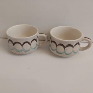 Könnte beinhalten: Zwei cremefarbene Keramik-Kaffeetassen mit einem braunen und türkisfarbenen Wellenmuster am Rand. Jede Tasse hat einen kleinen Henkel und eine gesprenkelte Oberfläche. Die Tassen stehen nebeneinander.