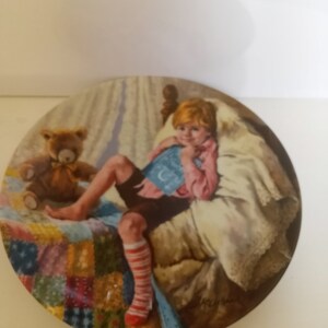 Könnte beinhalten: Ein runder Keramikteller mit einem bunten Patchwork-Steppdeckenmuster. Ein kleines Kind mit roten Haaren liegt im Bett und liest ein Buch. Ein Teddybär sitzt neben dem Kind auf dem Bett. Der Teller ist vom Künstler signiert.