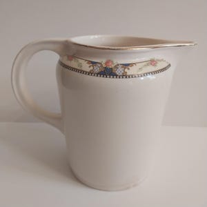 May include: A cream-coloured ceramic jug with a curved handle and a gold rim. The jug features a decorative band with pink roses, blue accents, and a black and gold border. A classic piece for serving beverages.
