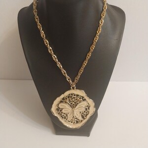 May include: Gold-tone necklace with a large pendant featuring a butterfly design. The pendant has a cream-colored butterfly in the center, surrounded by an openwork design and a cream-colored border. The chain is a twisted rope style.