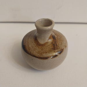 May include: A small, round, ceramic vase with a narrow neck. The vase is a light beige color with brown brushstrokes around the body. The vase has a smooth, matte finish and is suitable for small flowers or decorative purposes.