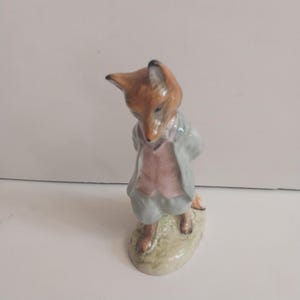 May include: A ceramic figurine of a fox wearing a light blue jacket and pink vest. The fox is standing on a small, oval-shaped base with green and yellow accents. The fox's fur is brown and its ears are black.