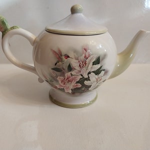 May include: A white ceramic teapot with a lid and a hummingbird-shaped handle. The teapot features a floral design with pink and white lilies and a hummingbird. The spout and base have a light green trim.