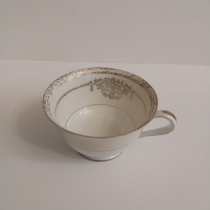 May include: A white teacup with a gold rim and handle. The cup features a decorative gold floral and leaf pattern around the rim. The cup is empty and sits on a white surface.