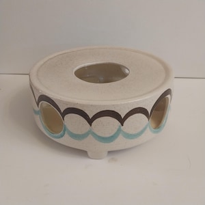 Może przedstawiać: Kremowy ceramiczny podgrzewacz do herbaty o okrągłym kształcie. Podgrzewacz posiada centralny otwór i jest ozdobiony brązowymi i turkusowymi wzorami fal na bokach. Ma trzy małe nóżki.