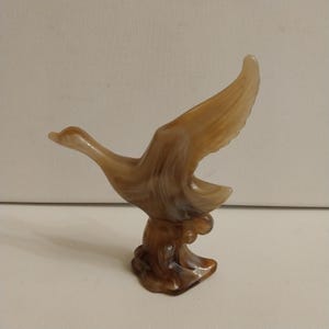 May include: A brown and tan glass figurine of a duck in flight. The duck is perched on a brown base, with its wings spread wide. The glass has a marbled appearance.
