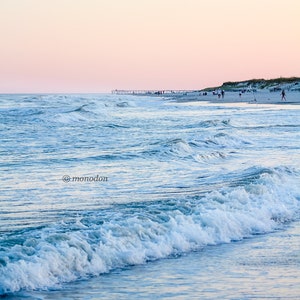 May include: A panoramic view of a beach with a pier in the distance. The sky is a soft pink and blue, and the waves are crashing on the shore.