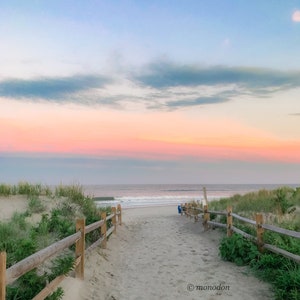 May include: A wooden fence leads to a sandy beach path with a view of the ocean and a pink and blue sunset sky.