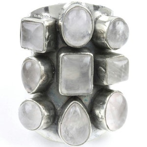 May include: A silver-toned ring featuring multiple light pink gemstones in various shapes, including squares, ovals, and teardrops. The gemstones are set closely together, creating a cluster design. The ring is set against a white background.