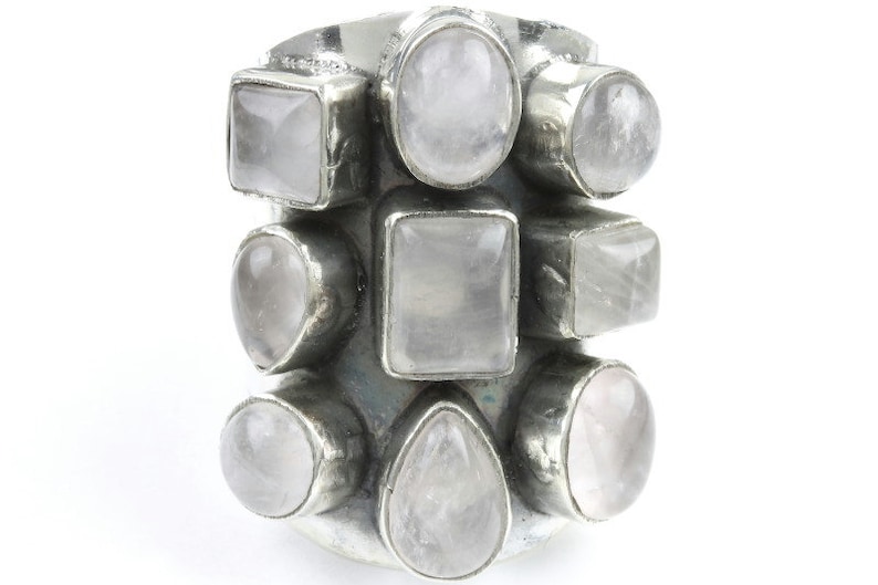 May include: A silver-toned ring featuring multiple light pink gemstones in various shapes, including squares, ovals, and teardrops. The gemstones are set closely together, creating a cluster design. The ring is set against a white background.
