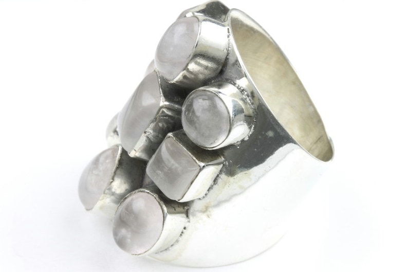 May include: A chunky silver ring featuring multiple light pink gemstones in various shapes. The ring has a wide band and the gemstones are set in individual silver bezels, creating a cluster design. The gemstones have a soft, translucent appearance.