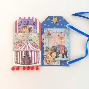 May include: A two-panel decorative tag featuring a circus theme. The left panel has a red and white striped tent and vintage ephemera. The right panel is blue with gold stars, a gold star embellishment, and an elephant with a performer.