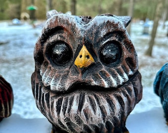 May include: A brown and white owl figurine with yellow beak and black eyes. The owl is sitting on a wooden surface covered in snow.