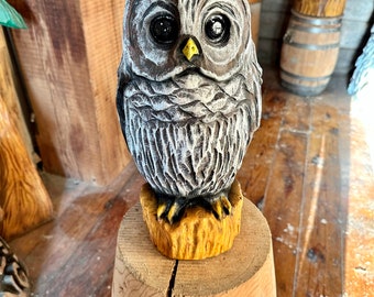 May include: A hand-carved wooden owl sculpture with a detailed, textured surface. The owl has large, dark eyes, a yellow beak, and is perched on a wooden base. The sculpture is painted in shades of gray and white, with a natural wood finish.