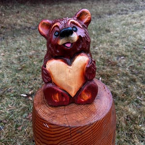 May include: A carved wooden bear figurine with a heart-shaped chest, sitting on a wooden stump. The bear is painted in shades of brown and tan, with a smiling expression. The heart is a lighter shade of wood. The stump is also made of wood.