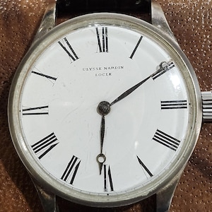 Puede incluir: Un reloj de pulsera plateado con una esfera blanca y marcadores de hora en n&uacute;meros romanos negros. El reloj tiene agujas negras y una correa de cuero negra. La esfera del reloj dice "ULYSSE NARDIN LOCLE".