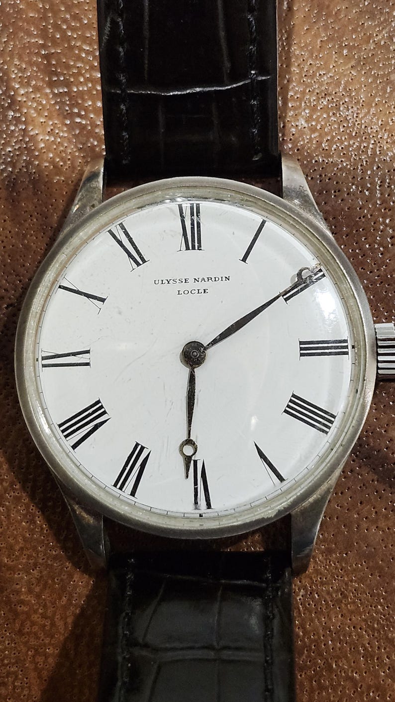 Puede incluir: Un reloj de pulsera plateado con una esfera blanca y marcadores de hora en n&uacute;meros romanos negros. El reloj tiene agujas negras y una correa de cuero negra. La esfera del reloj dice "ULYSSE NARDIN LOCLE".
