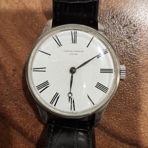 Puede incluir: Un reloj de pulsera plateado con una esfera blanca y marcadores de hora en n&uacute;meros romanos negros. El reloj tiene una correa de cuero negro y dice "Ulysse Nardin Locle" en la esfera.