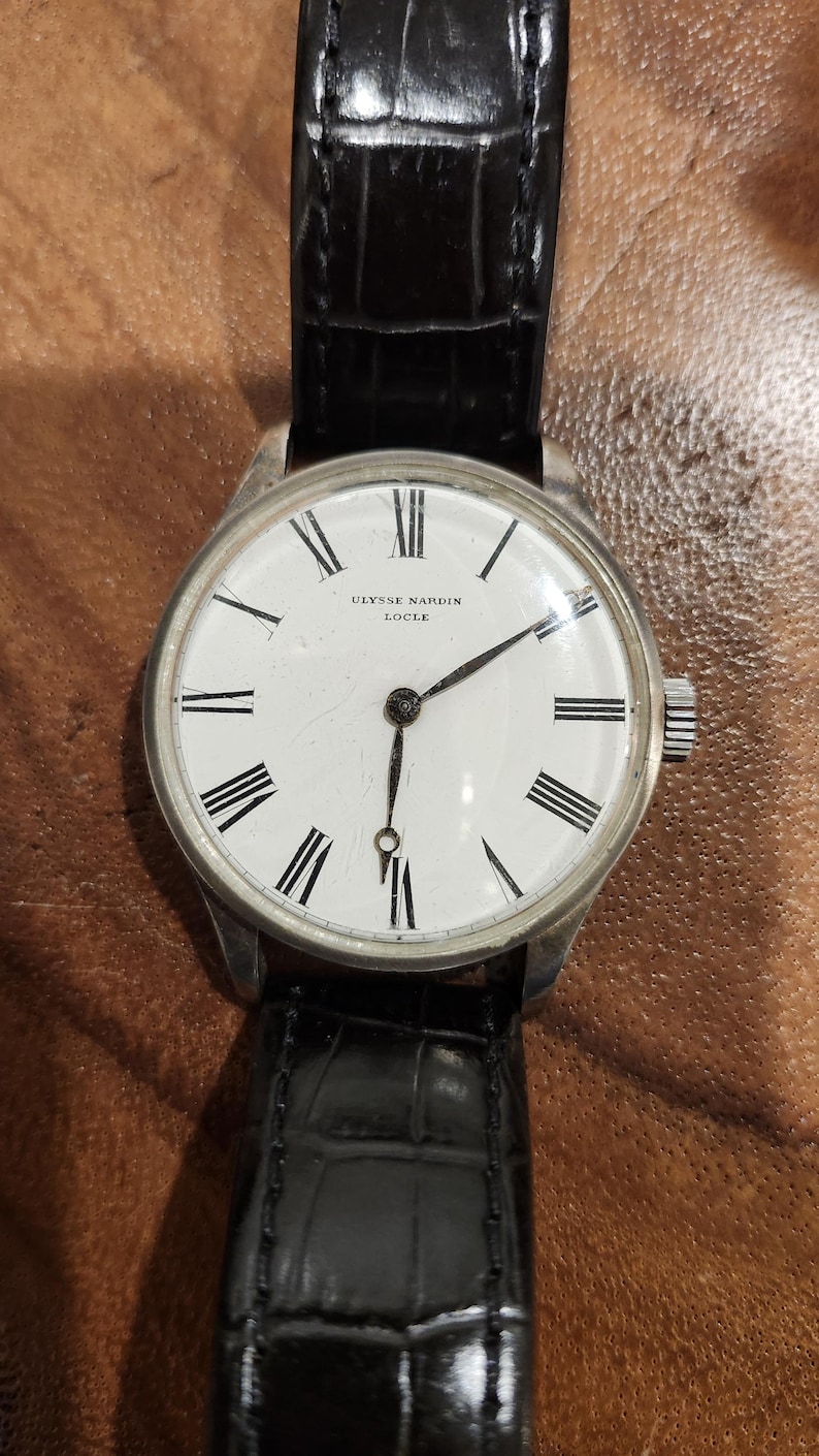 Puede incluir: Un reloj de pulsera plateado con una esfera blanca y marcadores de hora en n&uacute;meros romanos negros. El reloj tiene una correa de cuero negro y dice "Ulysse Nardin Locle" en la esfera.