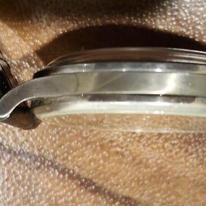 Puede incluir: Vista lateral de cerca de un reloj de pulsera plateado con una correa de cuero negro. El reloj tiene una esfera redonda y un acabado liso y pulido.