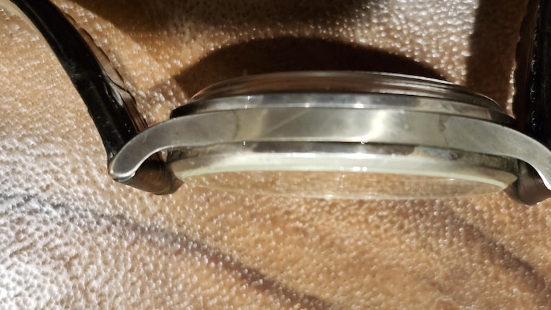Puede incluir: Vista lateral de cerca de un reloj de pulsera plateado con una correa de cuero negro. El reloj tiene una esfera redonda y un acabado liso y pulido.