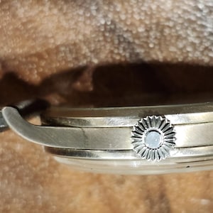 Puede incluir: Vista lateral de cerca de un reloj de pulsera plateado con una correa de cuero negro. El reloj tiene una esfera redonda y una corona decorativa.
